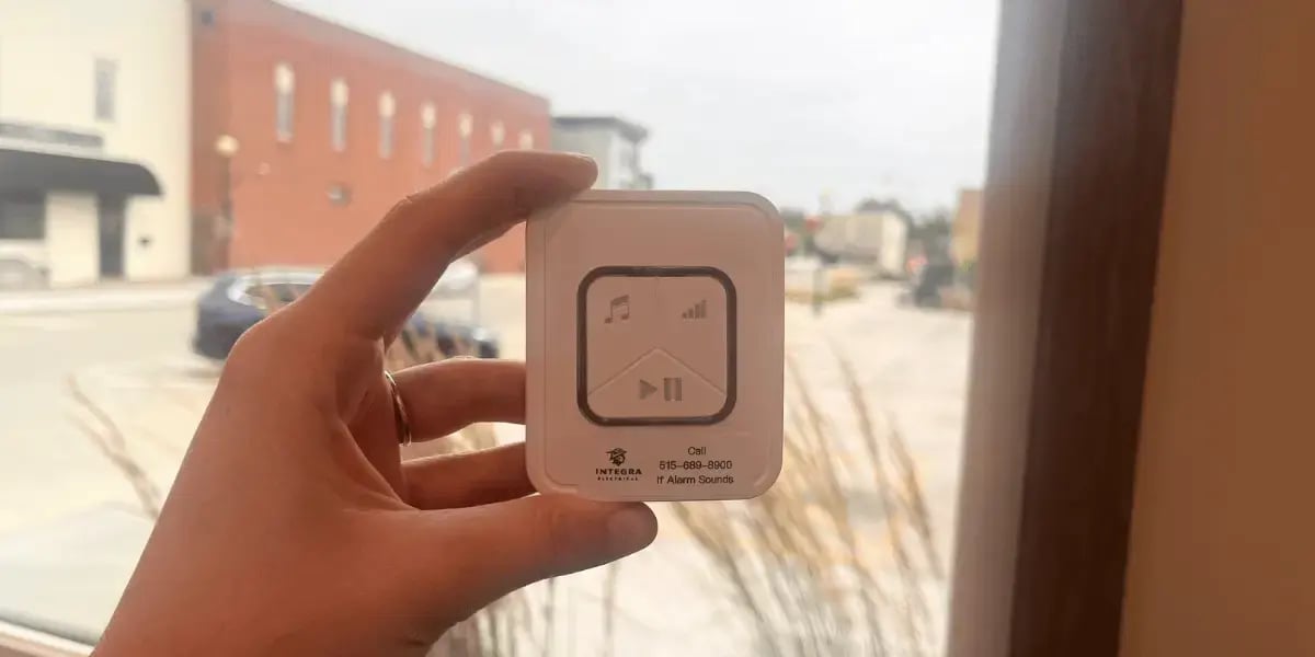 A hand holding a Bad Wiring Alarm device labeled with Integra Electrical’s contact number, against a small-town street view through a window.