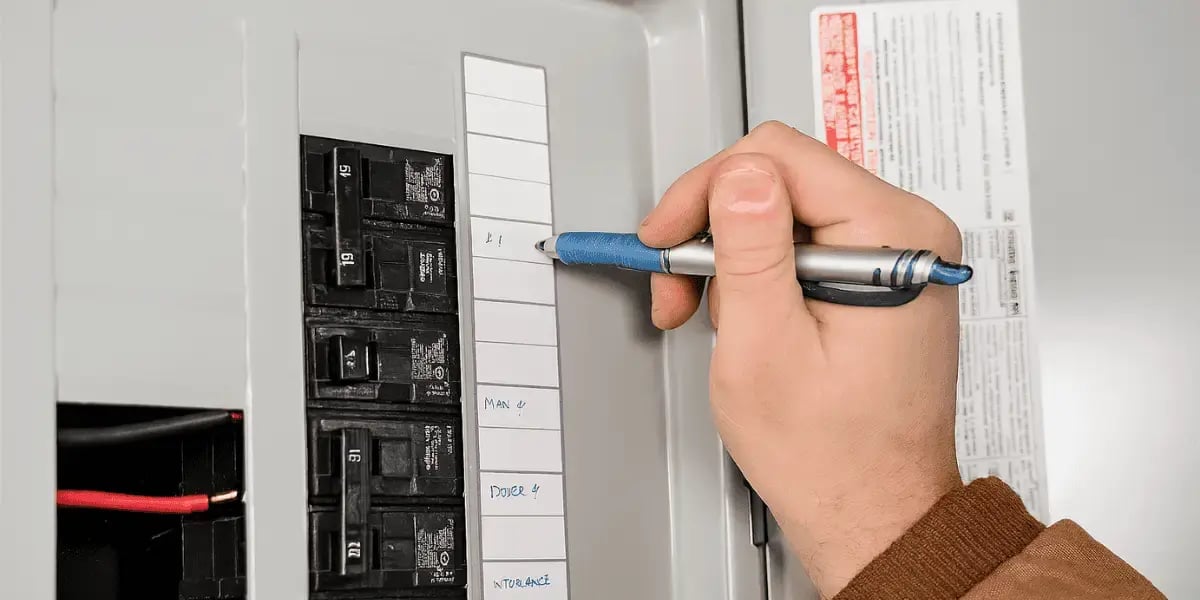 A close-up of an electrician labeling a residential breaker panel using a blue pen, with handwritten circuit names on a white panel chart and visible black circuit breakers arranged vertically.