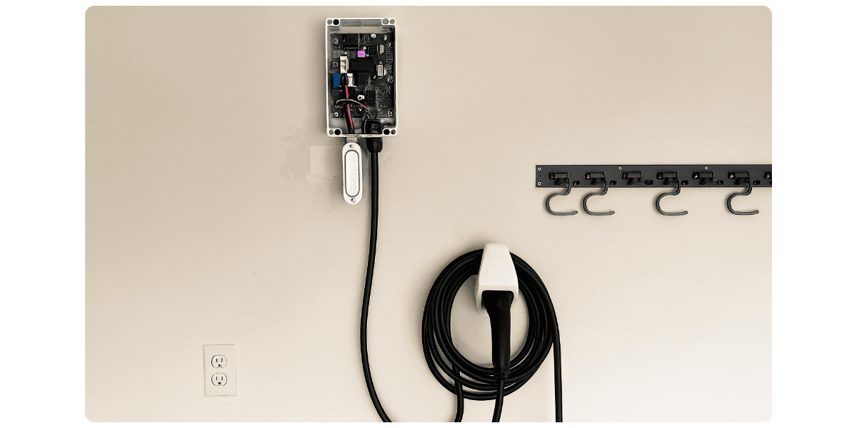 A wall-mounted EV charger installed in a garage, with the charging cable neatly coiled beside a row of empty wall hooks and an outlet below.