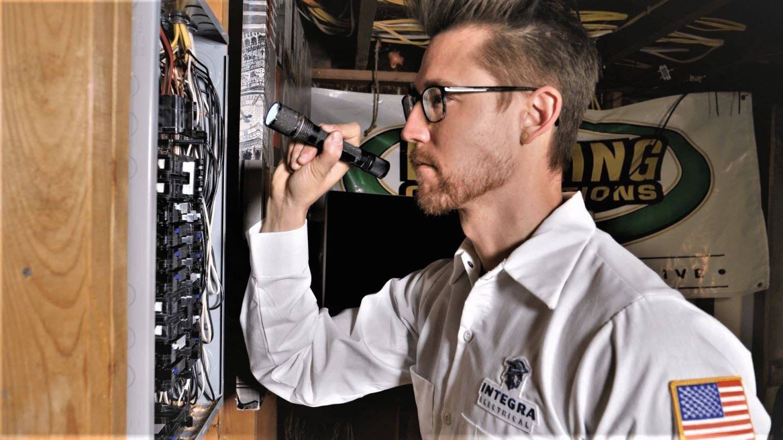 An electrician wearing a white Integra Electrical uniform uses a flashlight to inspect a residential electrical panel. The panel contains multiple breakers and wiring, and the setting appears to be a basement or utility area.