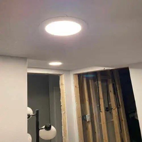 A ceiling with installed recessed lighting fixtures in a room under renovation. The lights are turned on, illuminating the space, which features exposed wooden framing and partially finished walls.