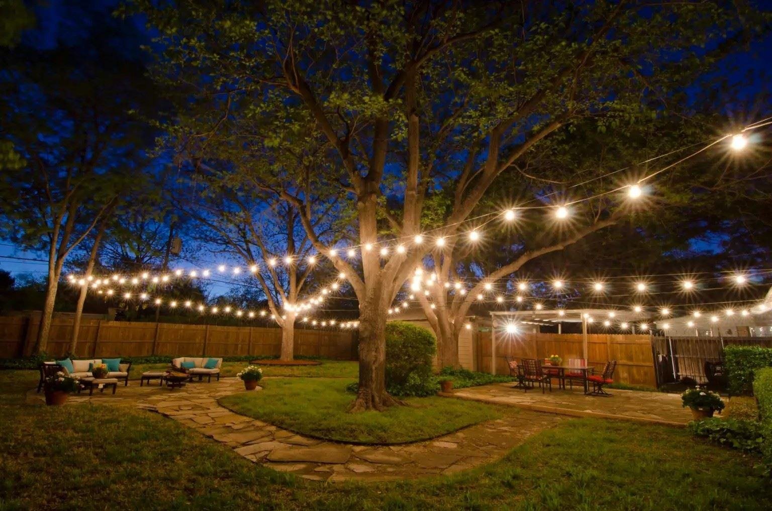A backyard at dusk lit with string lights hanging between large trees. The space includes a stone walkway, seating areas with chairs and cushions, a dining table, and a wooden privacy fence surrounding the yard.