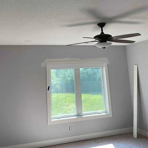 A ceiling fan is installed near the corner of a room, positioned directly above a window. The room has light gray walls, a large double-pane window, and a white baseboard along the floor.