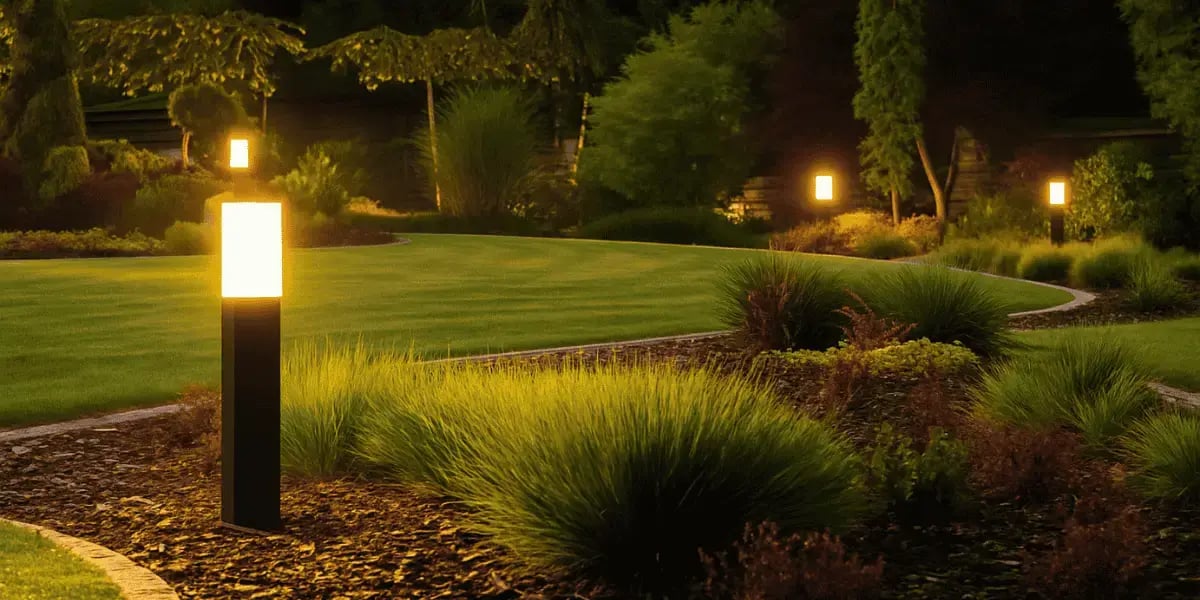 A beautifully landscaped backyard at dusk, featuring modern black bollard lights casting a warm glow along a curved mulch path bordered by manicured grass, shrubs, and ornamental trees.