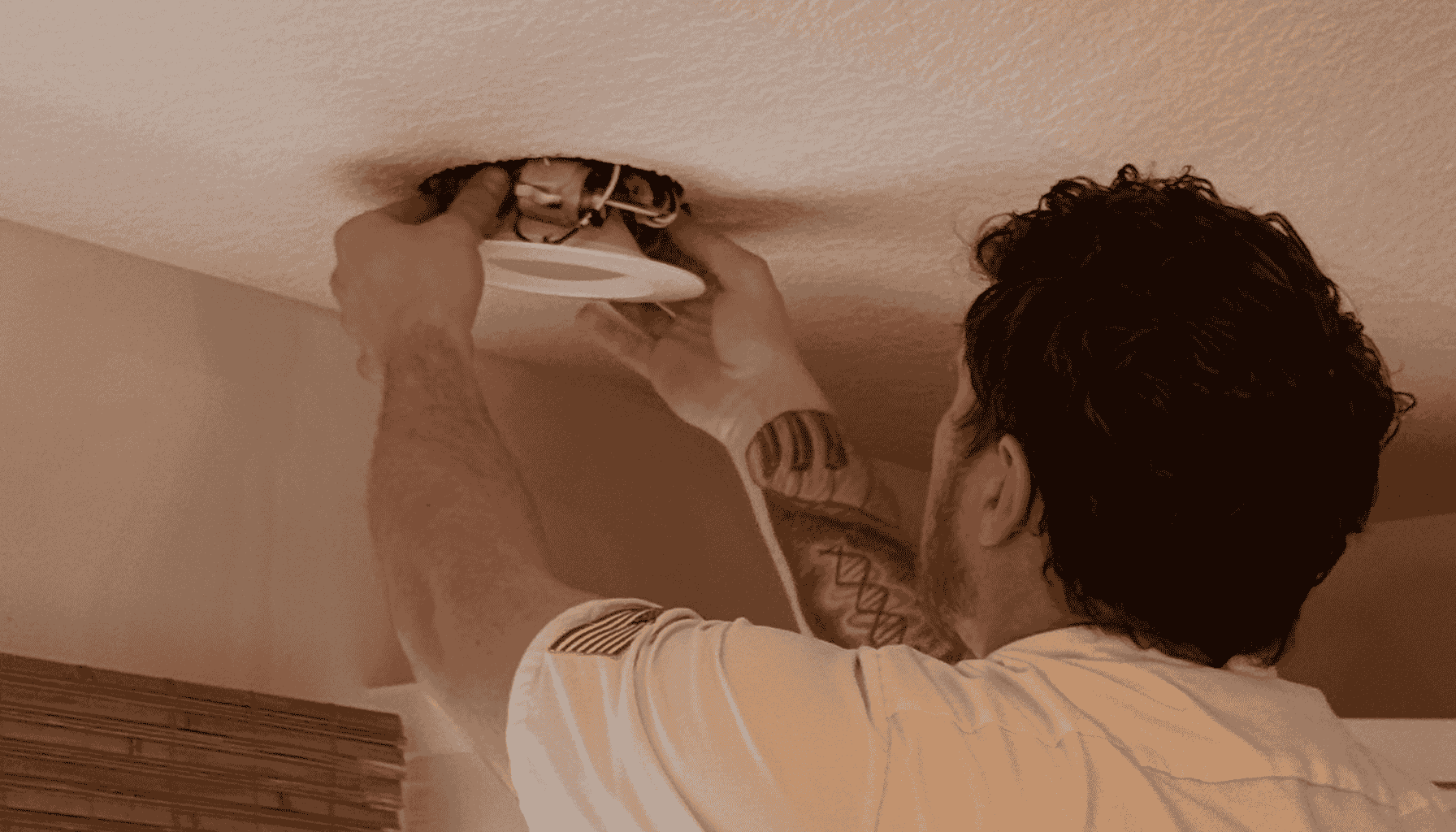 Electrician installing a recessed light fixture into a ceiling, with exposed wires visible and wearing a white uniform shirt.