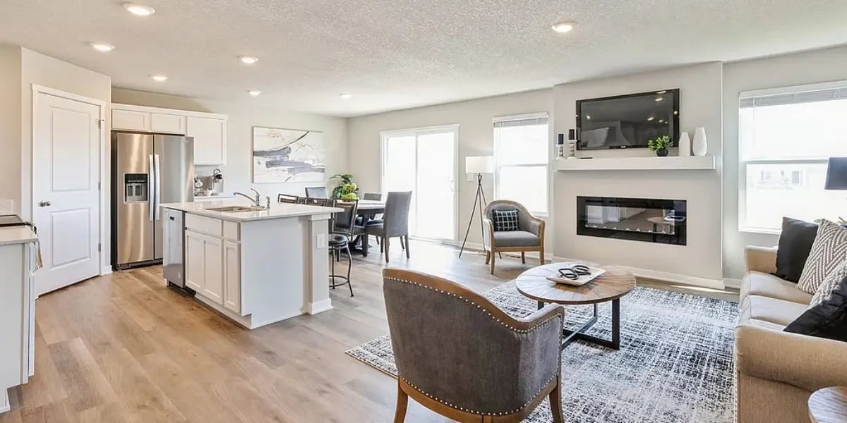 Modern open-concept kitchen and living room with recessed lighting, stainless steel appliances, neutral decor, and a wall-mounted fireplace beneath a flat-screen TV.