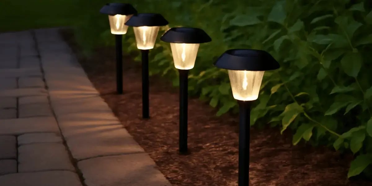A row of four solar-powered outdoor path lights glows softly along a stone walkway at dusk, bordered by mulch and lush green plants.