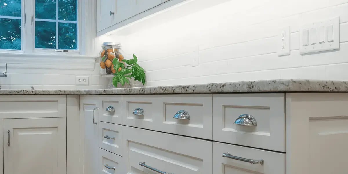 A bright, modern kitchen with white Shaker-style cabinets, speckled granite countertops, and sleek under-cabinet LED lighting, featuring a jar of fruit and a small potted plant near the sink under a large window.