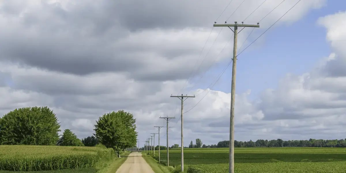 A wide landscape view of a straight gravel road running through lush green Iowa farmland. Telephone poles line the right side of the road, stretching into the distance. Corn and soybean fields border both sides under a bright sky filled with large, partly cloudy formations.
