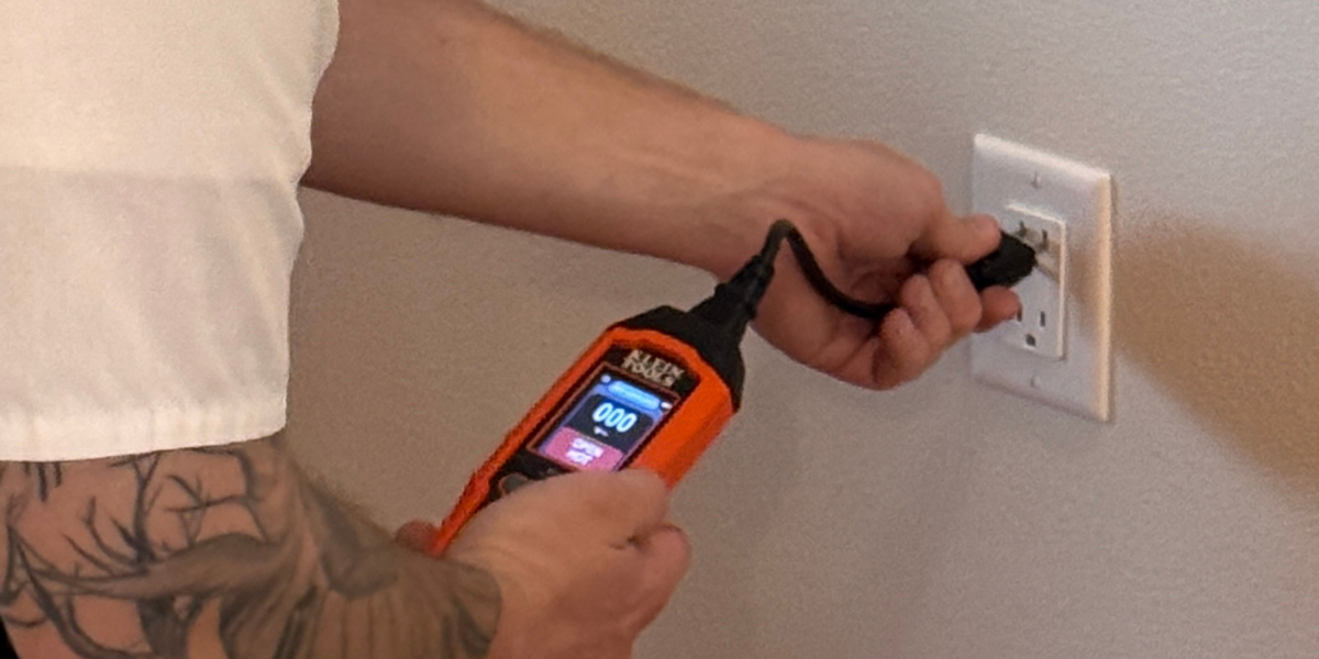 Close-up of an electrician using an orange voltage tester to check a wall outlet for voltage loss.