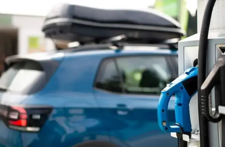 An electric vehicle charging station with a blue charging connector plugged into the unit. A blue SUV with a rooftop cargo box is parked nearby, slightly out of focus in the background.