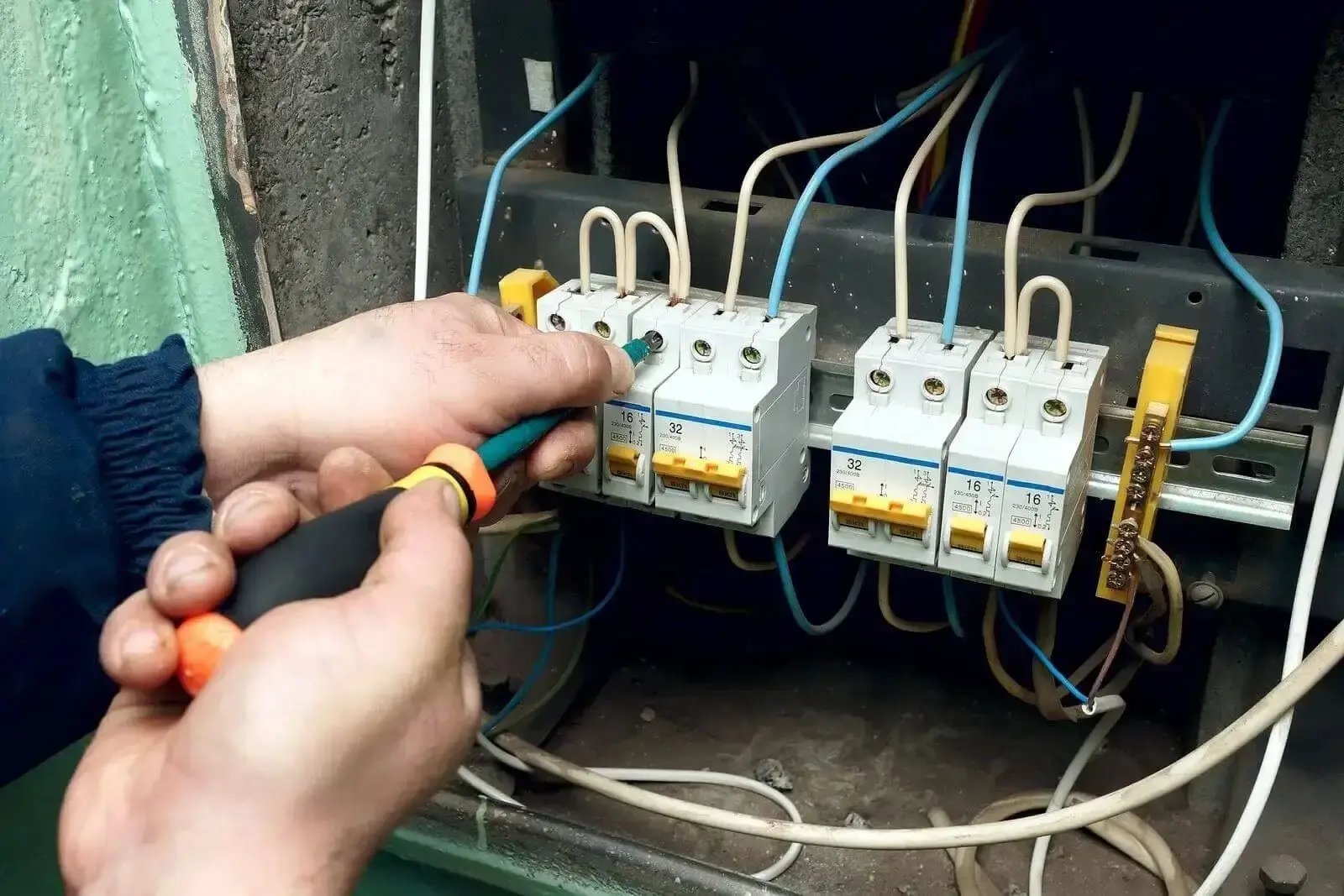 A person uses a screwdriver to work on a set of circuit breakers inside an open electrical panel. Multiple wires are connected to the breakers, and the setup includes white and blue insulated wires and yellow switch levers.