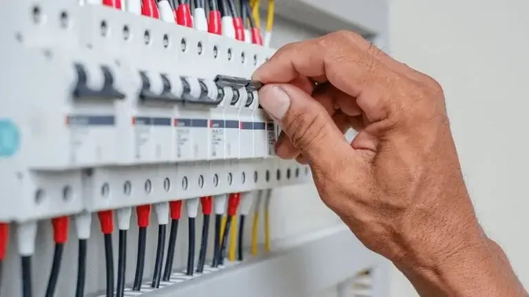 A close-up of a person’s hand flipping a circuit breaker switch inside an electrical panel. The panel contains multiple breakers connected to black, red, yellow, and white wires.