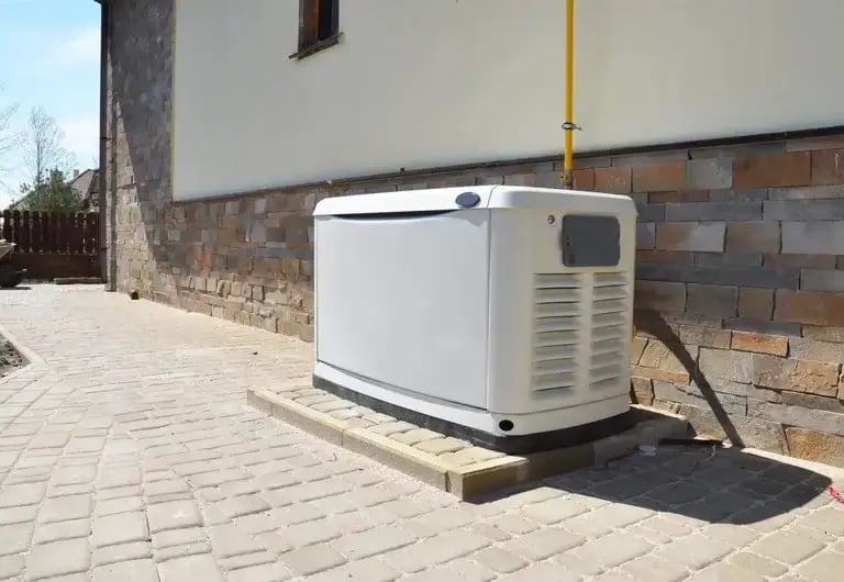 A standby generator installed on a concrete pad next to a residential building with a brick and stone exterior. A yellow gas line is connected to the unit, which is positioned on a paved walkway.