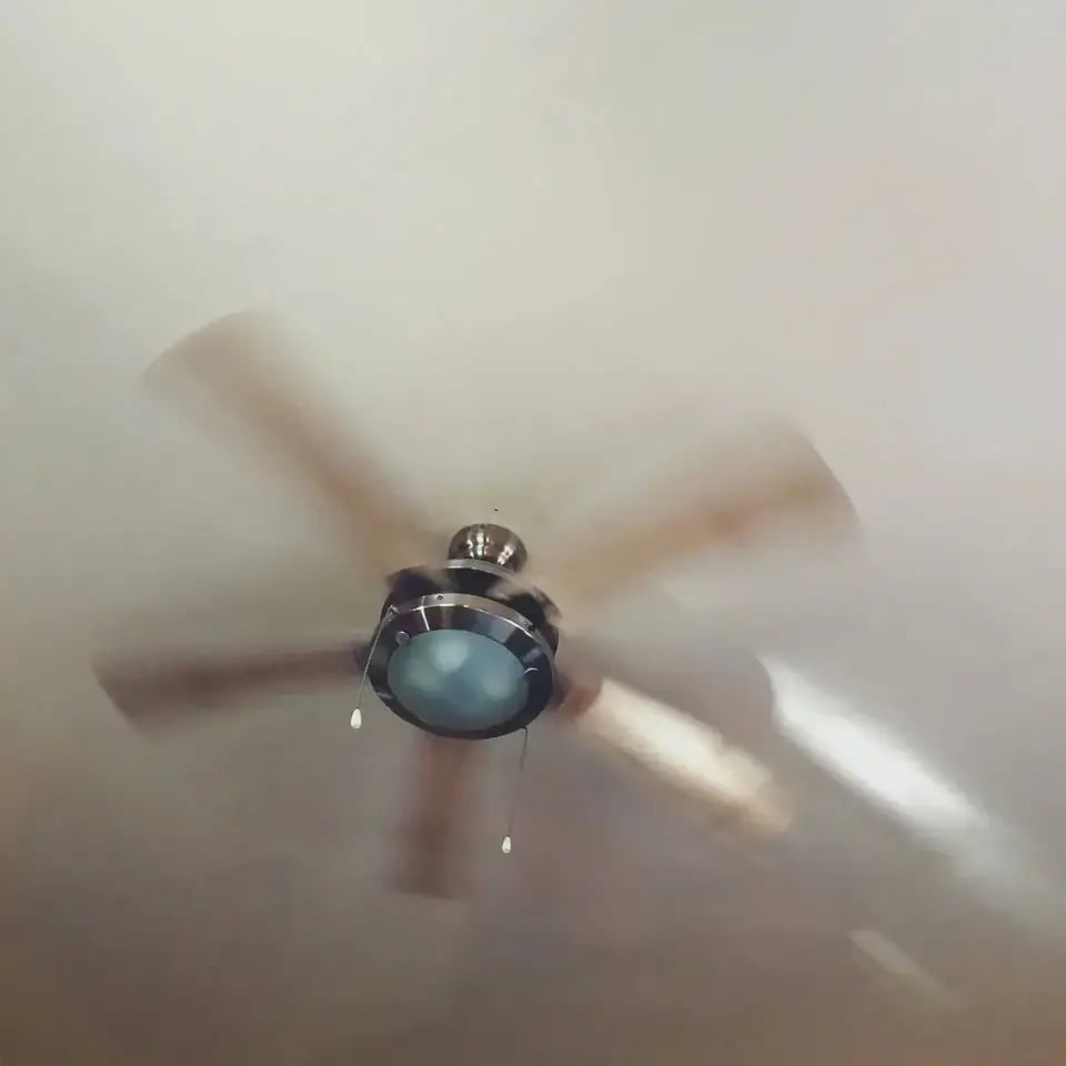 A ceiling fan with a built-in light fixture is spinning, creating a motion blur effect on its blades. The fan has a modern design with pull chains and is mounted on a light-colored ceiling.