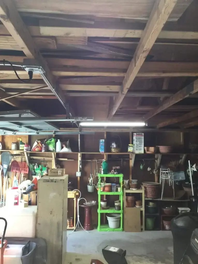 Fluorescent light fixture installed in a garage with exposed wooden beams, shelves, and stored tools and supplies.