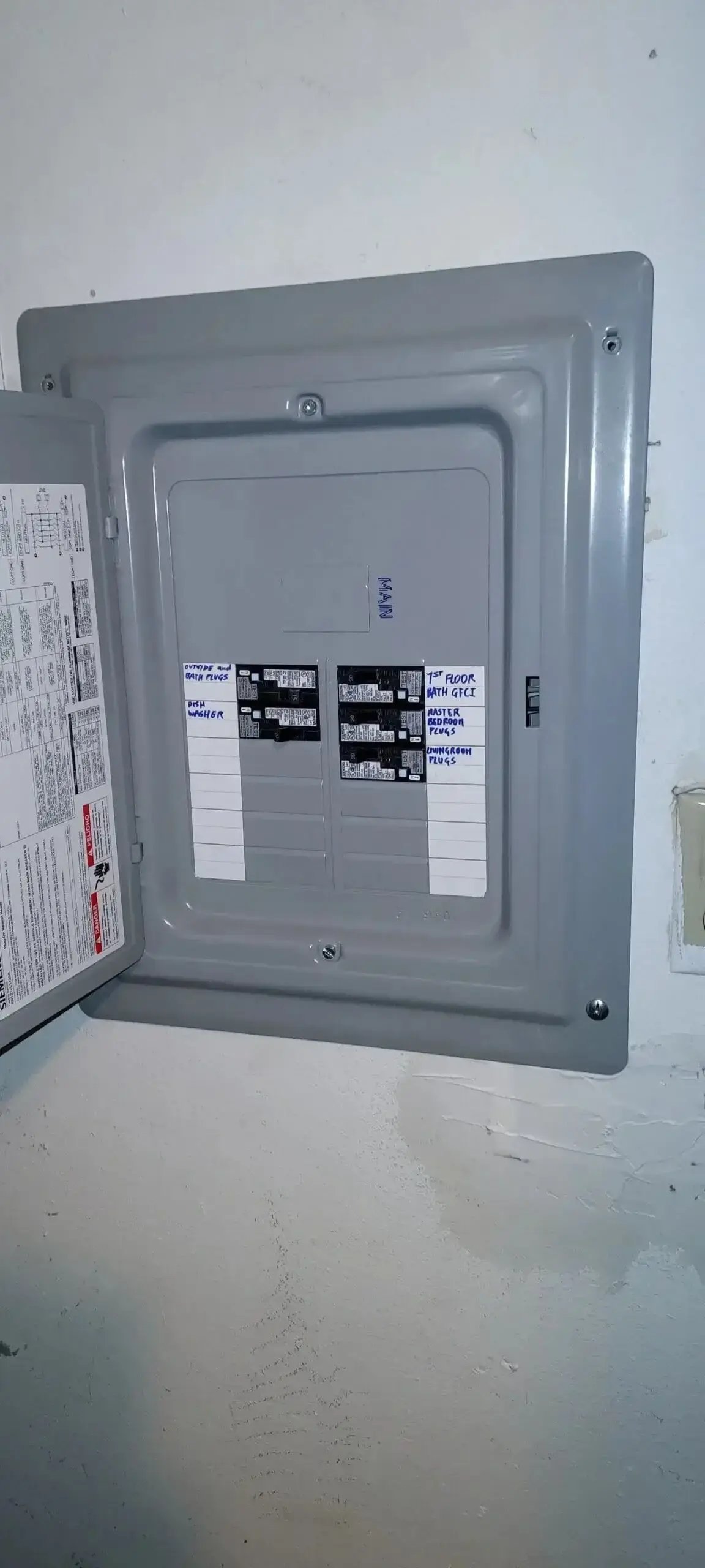 A residential electrical panel with the door open, showing labeled circuit breakers. Handwritten labels on the panel indicate specific circuits for areas like the bathroom, master bedroom, living room, and dishwasher.