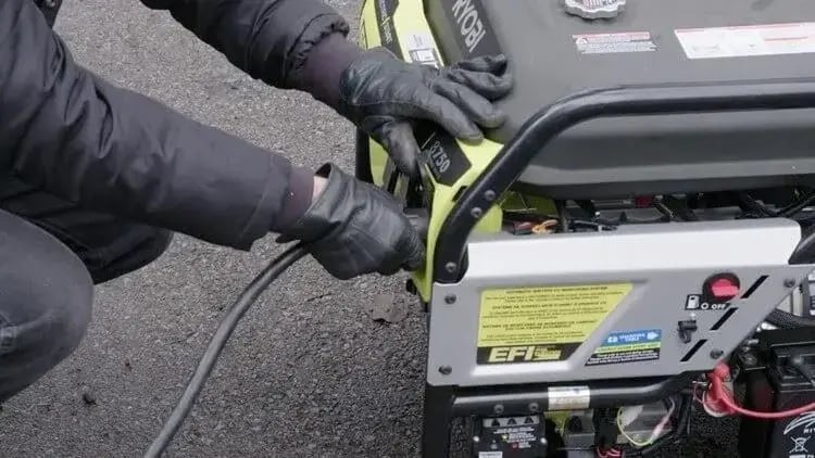 A person wearing black gloves connects a heavy-duty power cable to a portable generator on an asphalt surface. The generator features a control panel with labels and warning stickers, and the individual appears to be preparing it for use.