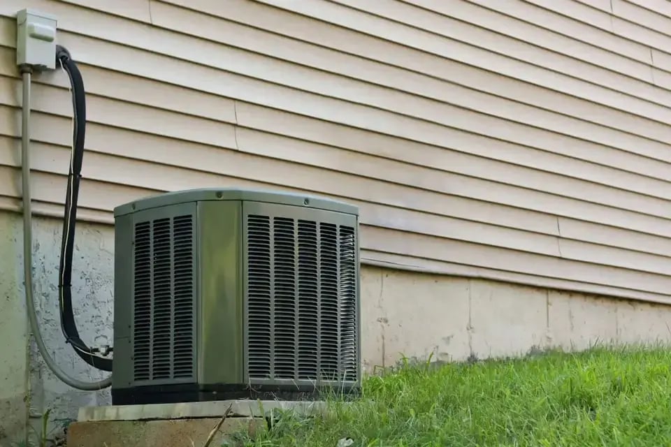 Outdoor air conditioning unit installed next to a beige house exterior with grass in the foreground.