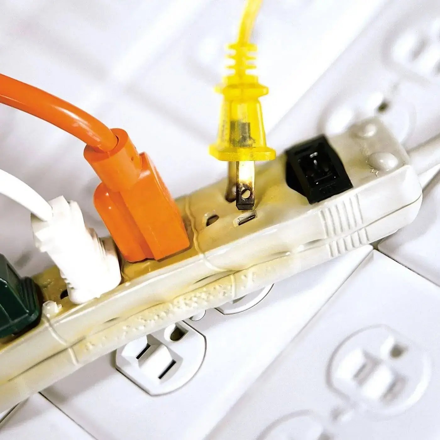 A close-up image of a power strip with multiple cords plugged in, showing signs of overheating and discoloration. The plastic casing appears warped and yellowed, indicating potential electrical damage or fire risk.