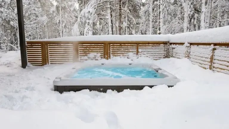 An outdoor hot tub with bubbling water sits surrounded by deep snow in a fenced area, with snow-covered trees and a wooden privacy fence in the background. Steam rises from the water into the cold winter air.