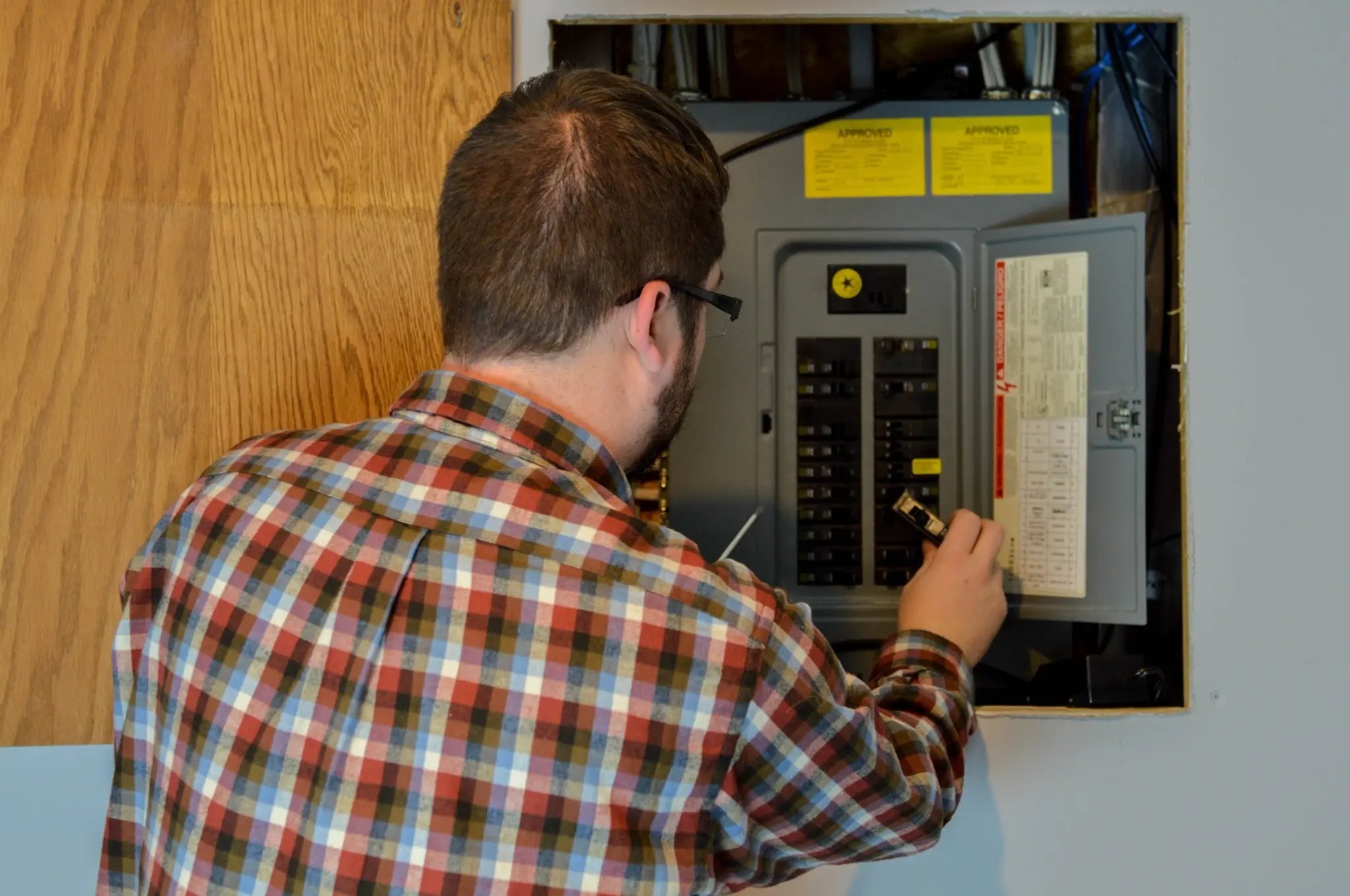 An electrician in a plaid shirt inspecting a breaker panel
