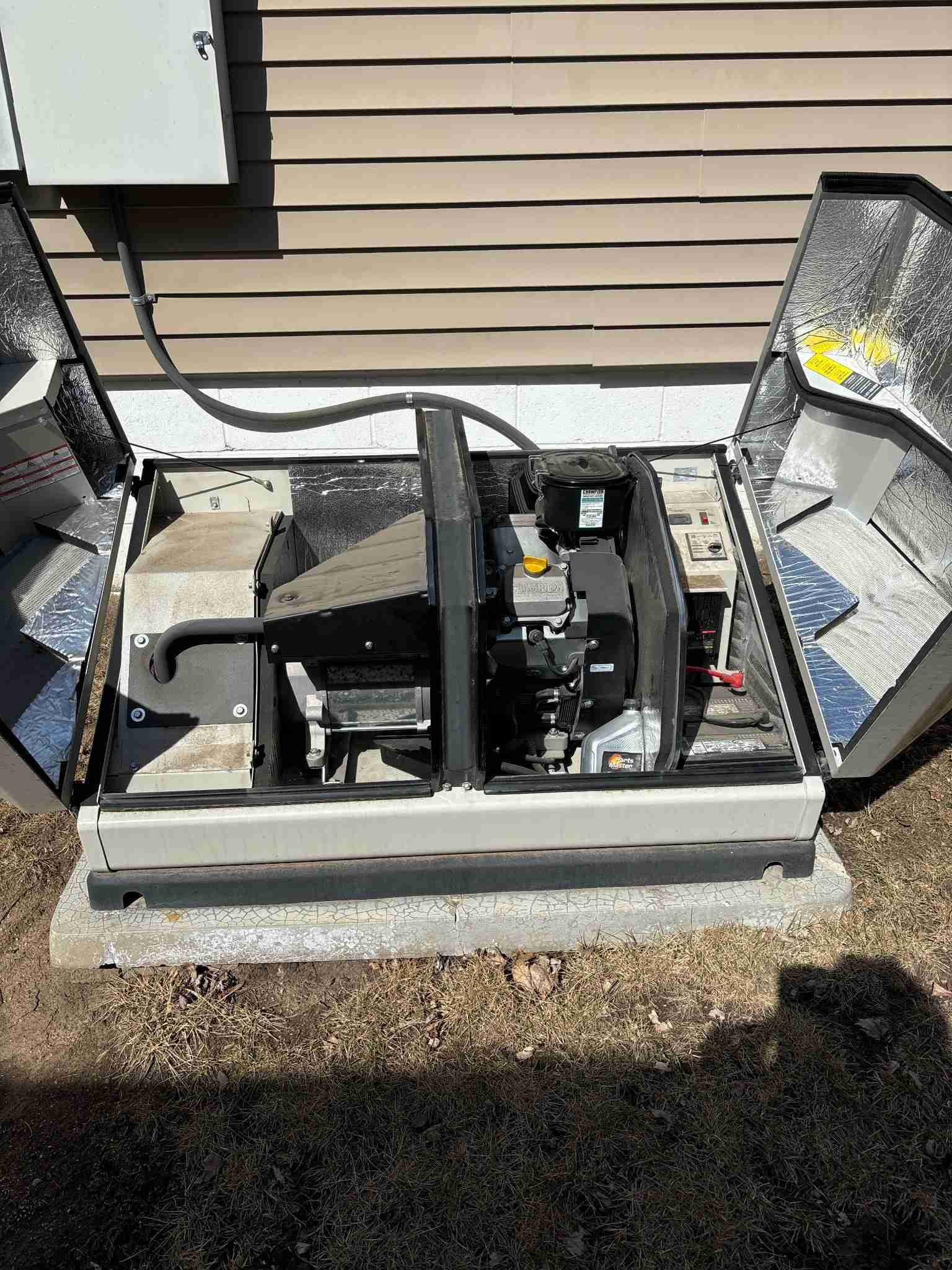 An open standby generator installed on a concrete pad outside a house, with its side panels removed to reveal the internal engine and components. The house exterior has beige vinyl siding and a nearby electrical conduit runs into a mounted box.