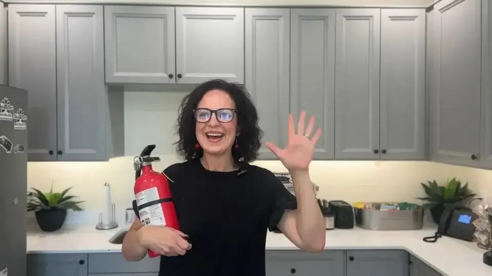 Person smiling and waving while holding a red fire extinguisher in a modern kitchen.