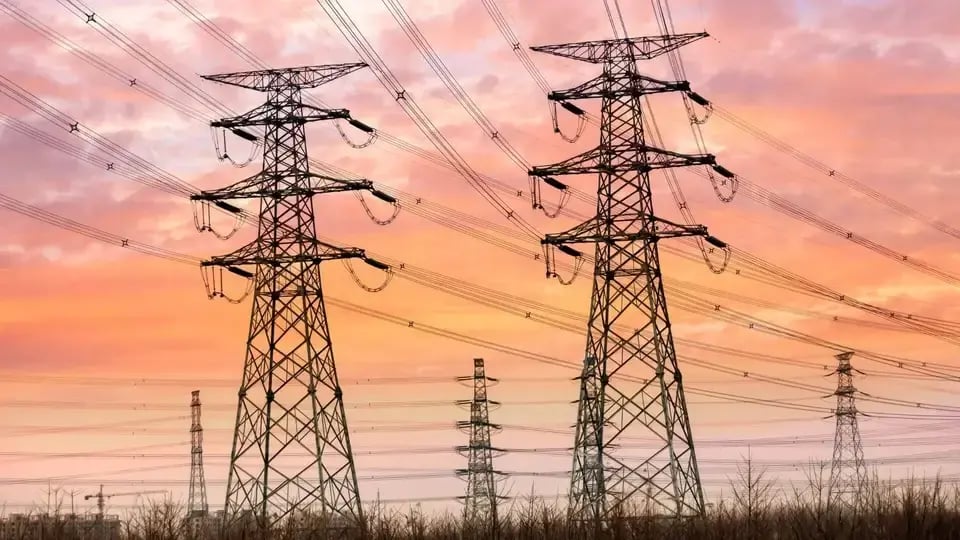 High-voltage transmission towers with multiple power lines stretching across the landscape at sunset, with an orange and pink sky in the background.