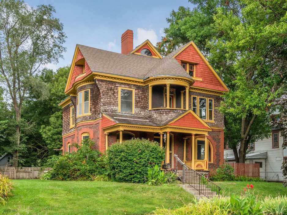 A large, two-story Victorian-style house with ornate architectural details, including a rounded turret, gabled rooflines, and a covered front porch. The exterior features a mix of red brick, wood shingles, and yellow trim, surrounded by trees and a well-kept lawn.