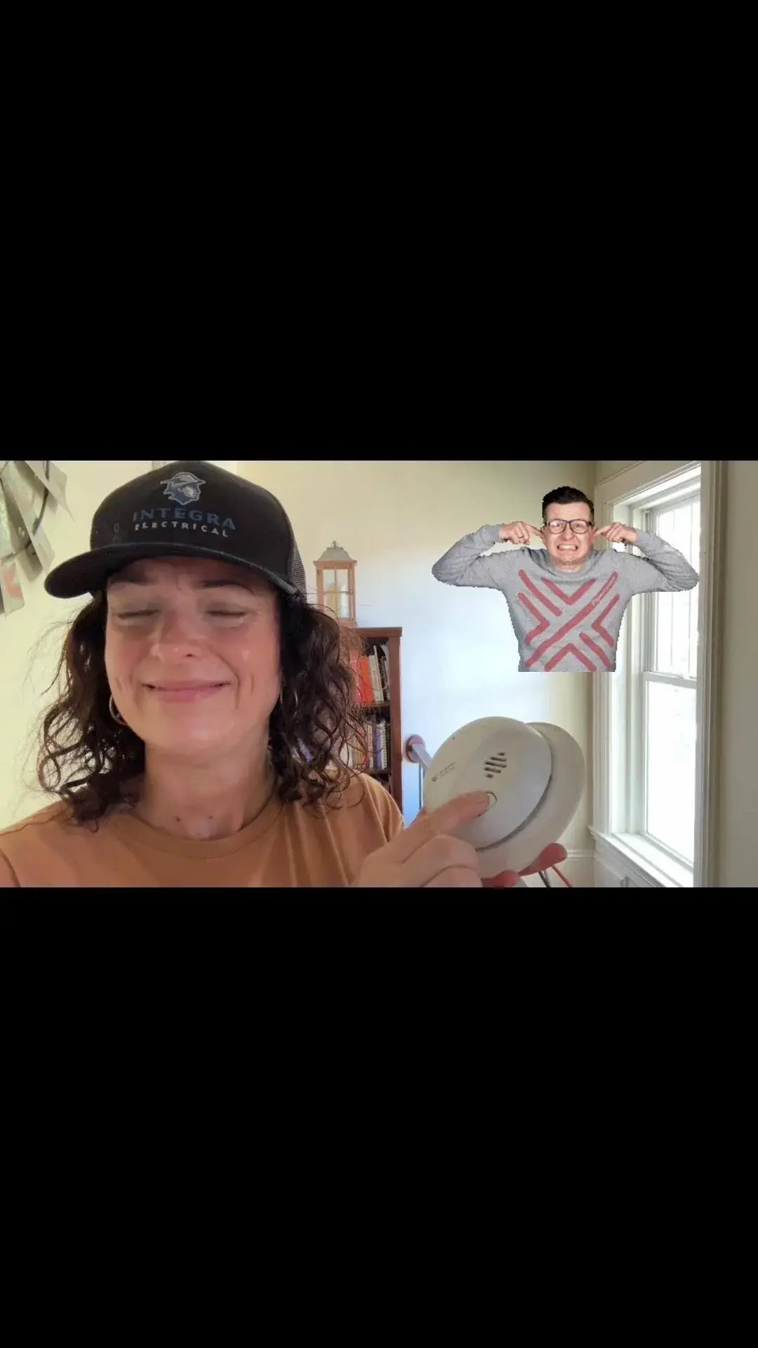 A woman wearing an Integra Electrical hat smiles while pressing the test button on a smoke detector, with a man in the background covering his ears in playful reaction to the alarm sound.