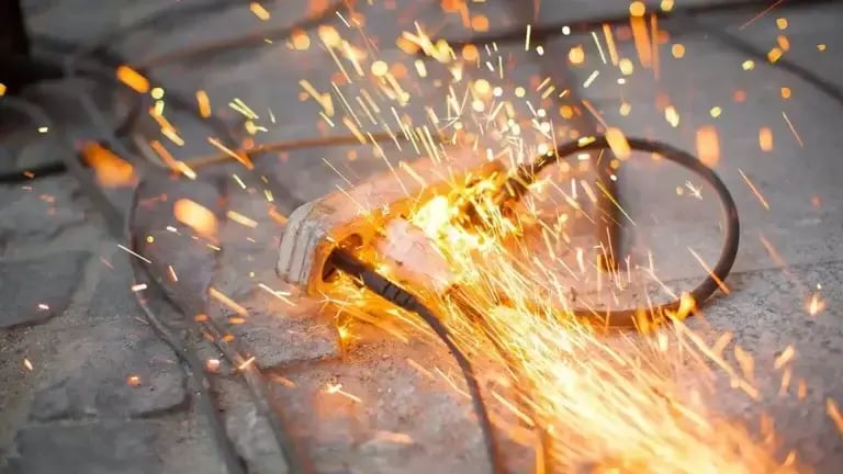 An electrical power strip on a concrete surface is sparking and catching fire, with bright orange sparks flying in multiple directions. Multiple cords are plugged into the strip, indicating a possible overload or short circuit.