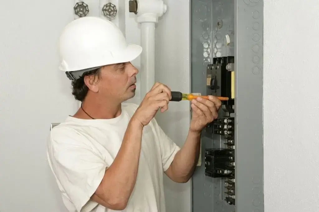 A person wearing a white hard hat and t-shirt uses a screwdriver to work on a residential electrical panel. The panel is open, revealing multiple breakers and internal wiring.